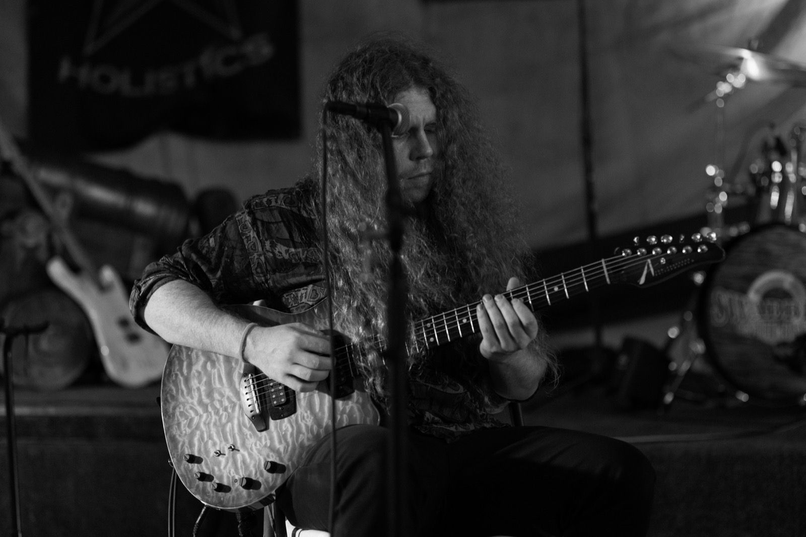 A guitarist with long curly hair plays a quilted-top electric guitar under a single stage light.